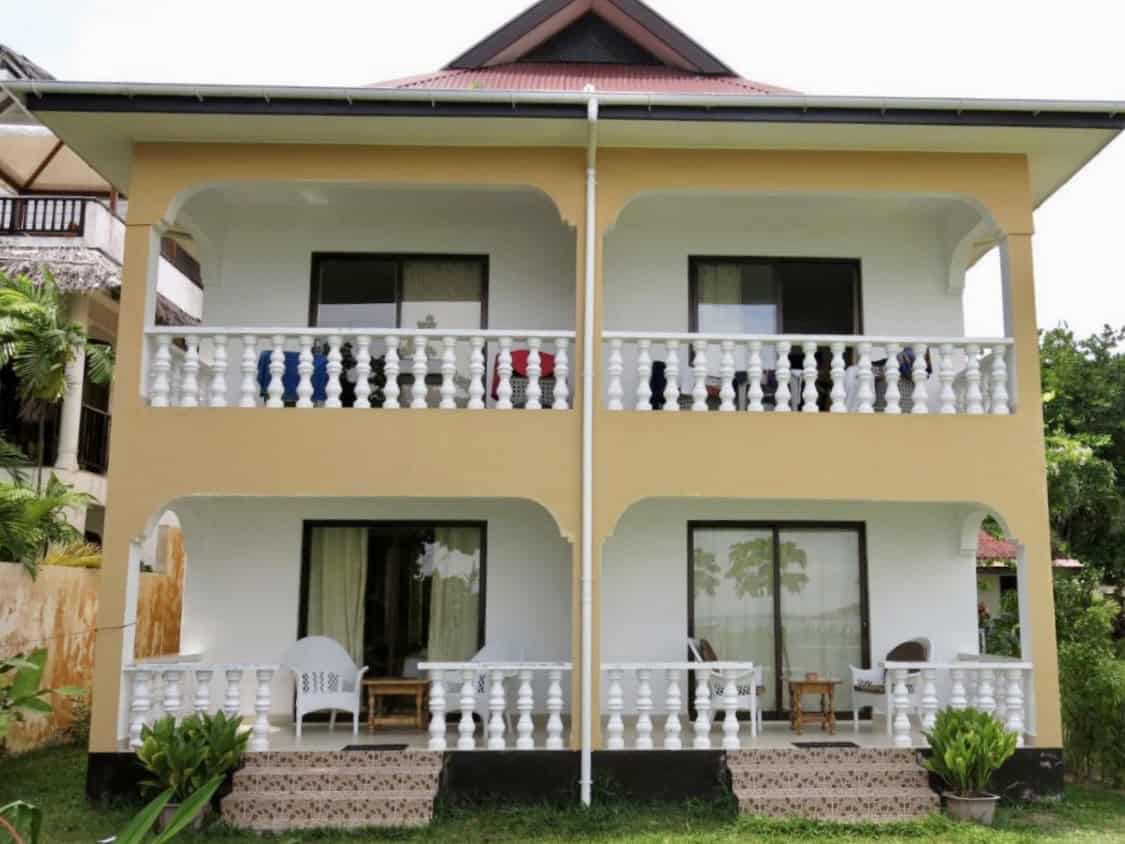 Balcon d'une maison à deux étages, maison colorée à Seychelles, maison avec balustrades blanches, maison avec terrasse extérieure et mobilier de patio, maison de vacances tropicale, architecture résidentielle seychelloise, balcon avec vue sur le jardin, maison avec railing décoratif, habitation de style créole, résidence avec espace extérieur, structure résidentielle avec mezzanine.