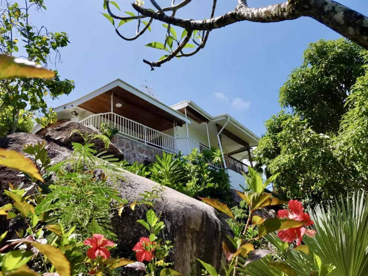 Vouette de maison typique seychelloise entourée de végétation luxuriante et de corail, vue depuis le sol avec un ciel bleu clair.