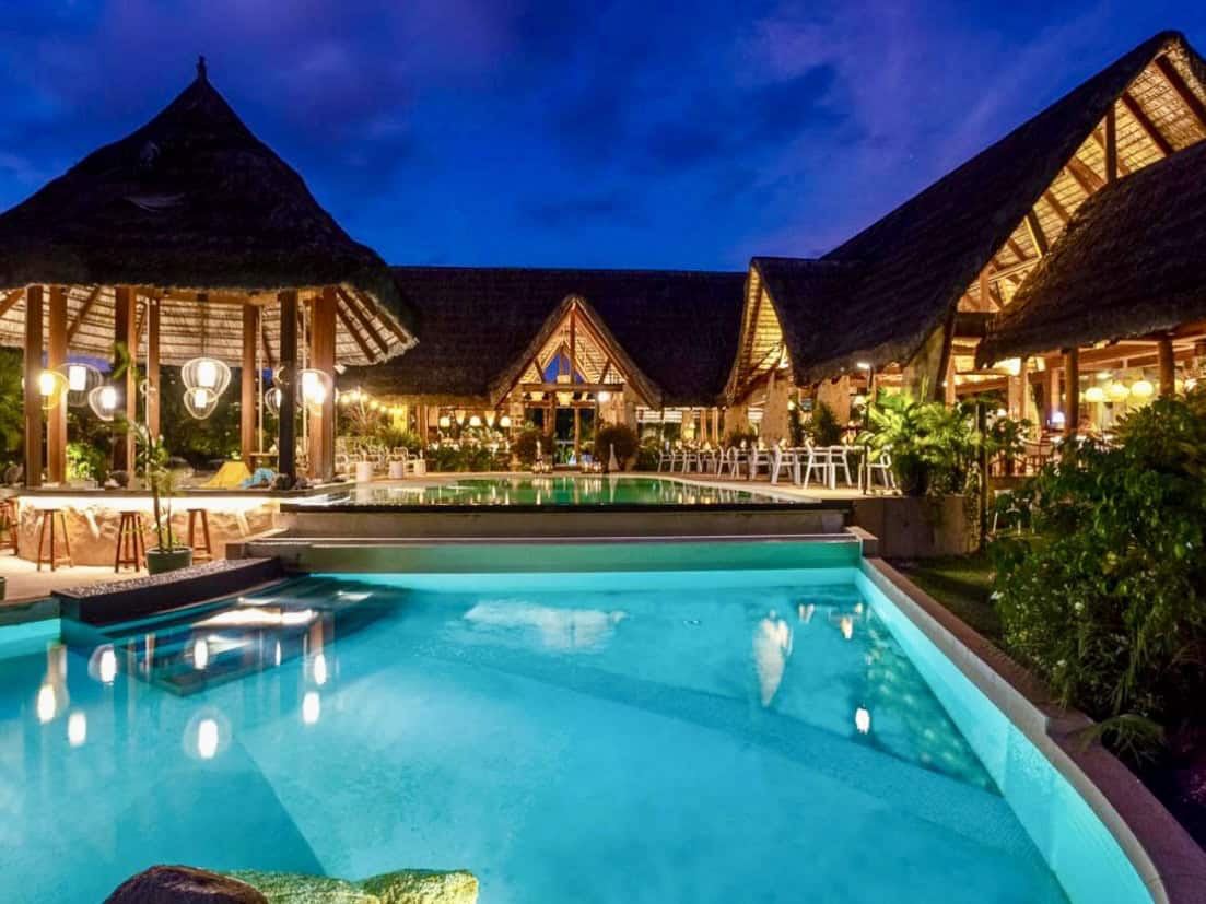Piscine éclairée et restaurant avec toiture en coques en chaume à Mahé, Seychelles, silhouette de jungle la nuit.