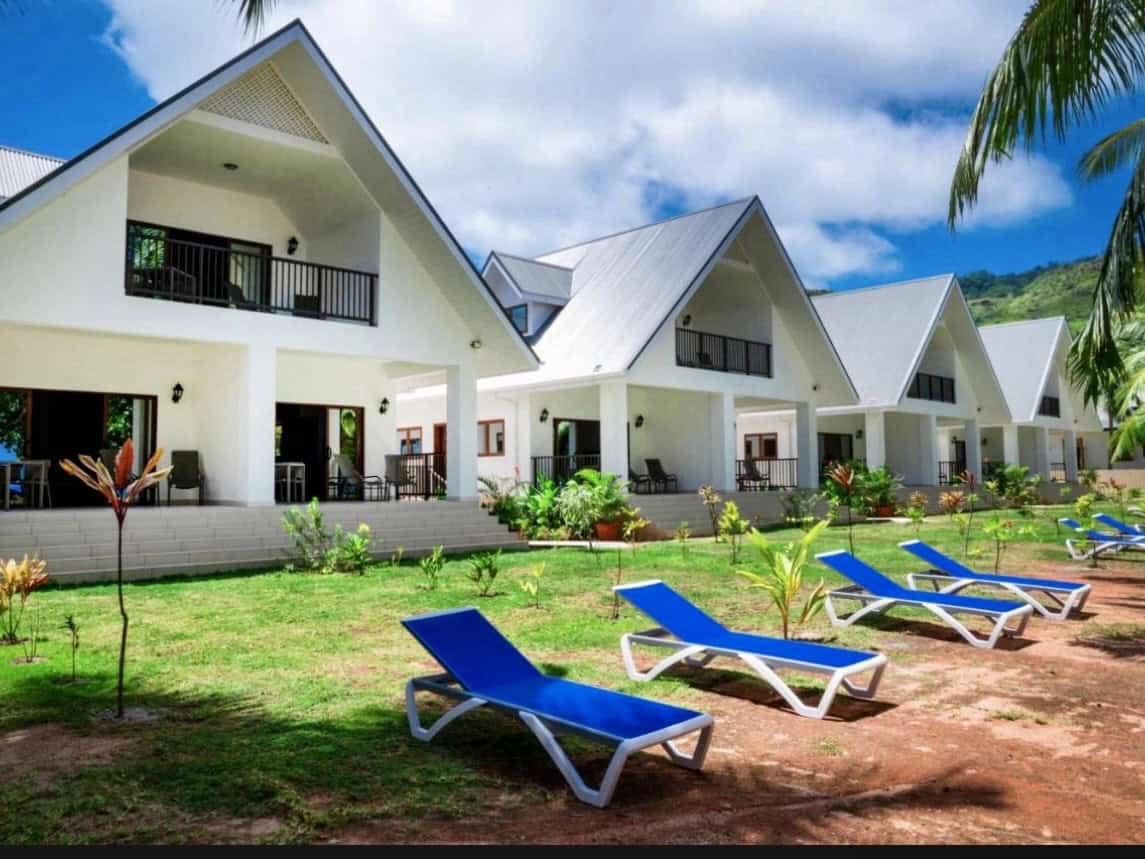 Modern white bungalows with sloped roofs on lush green grass, featuring outdoor lounge chairs, tropical plants, and a clear blue sky, ideal for a Seychelles vacation.