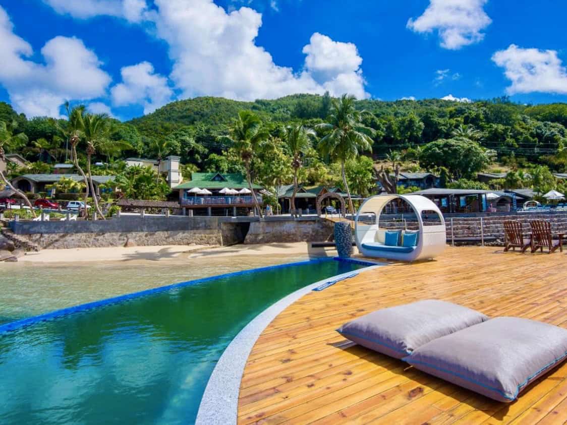 Bassin à débordement avec vue sur la mer turquoise et paisible, entouré de bois chaleureux et de coussins confortables, face à une baie tropicale verdoyante sous un ciel ensoleillé.