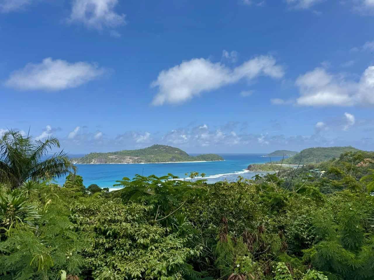 Plage turquoise sous ciel bleu à Seychelles, végétation luxuriante, îles verdoyantes en arrière-plan, paysage tropical, paradis côtier, nature intacte, vacances de rêve, environnement naturel, beauté sauvage, destination touristique, horizon maritime, vues panoramiques.