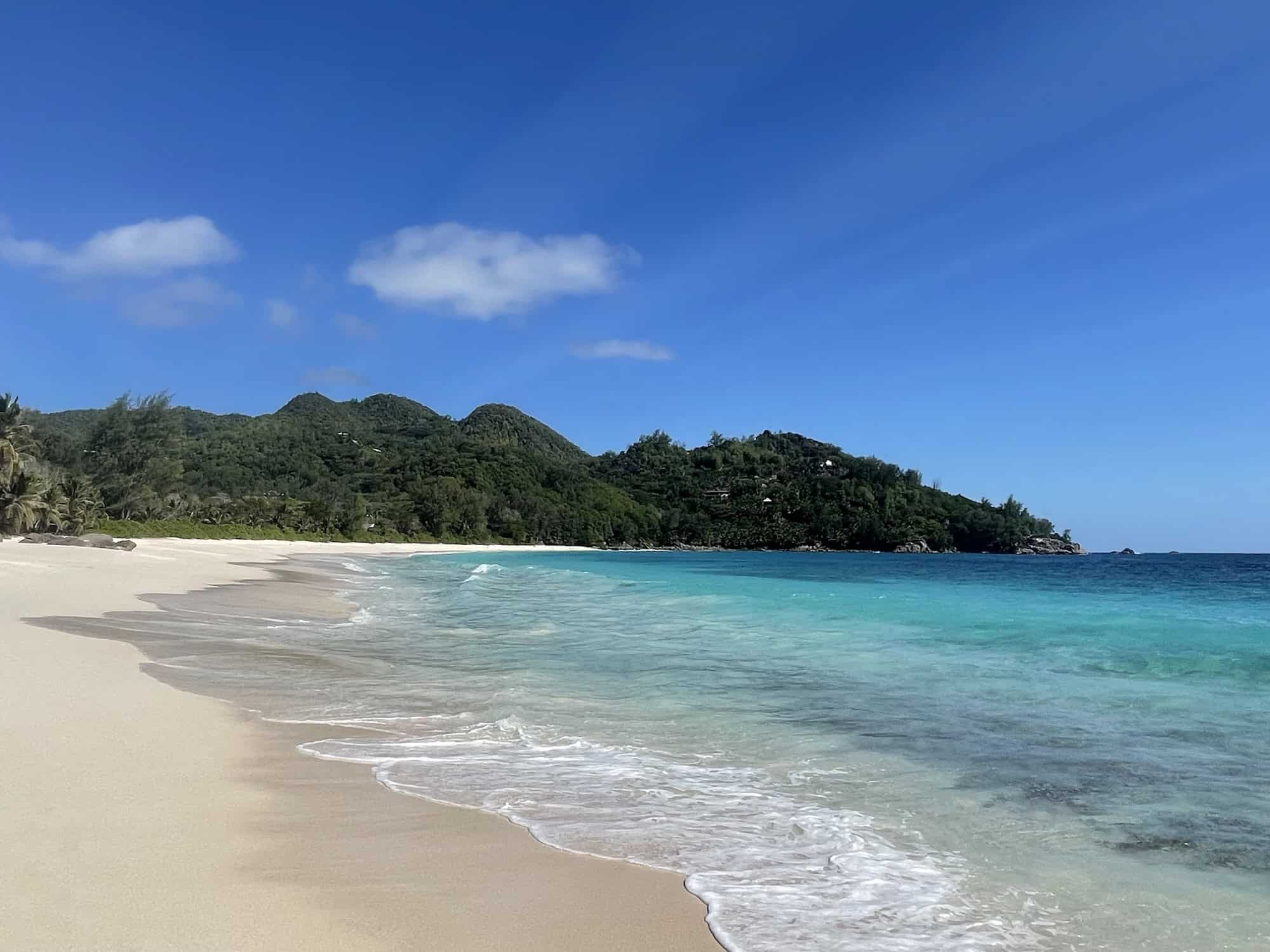 Plage de sable blanc avec vue sur mer turquoise et montagnes verdoyantes, typique des Seychelles.